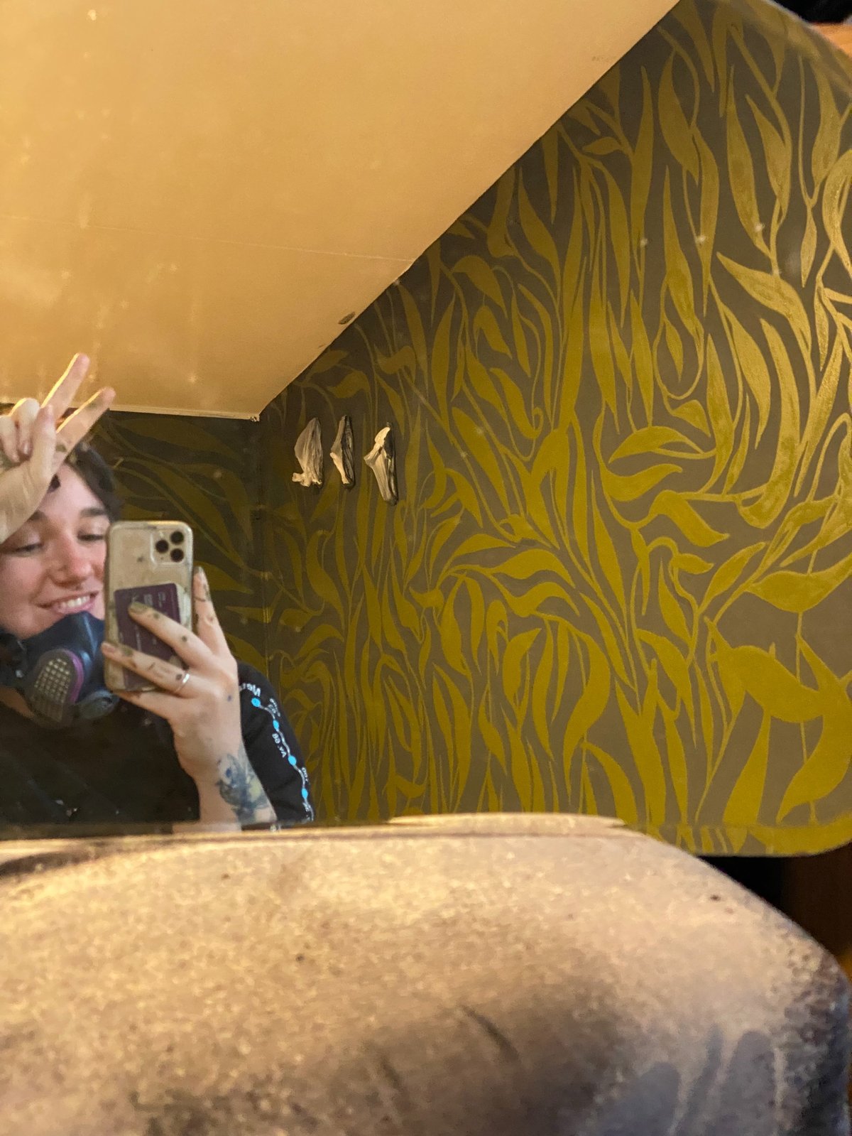 Person taking a mirror selfie in a room with gold and dark striped wallpaper, holding a phone up to their face