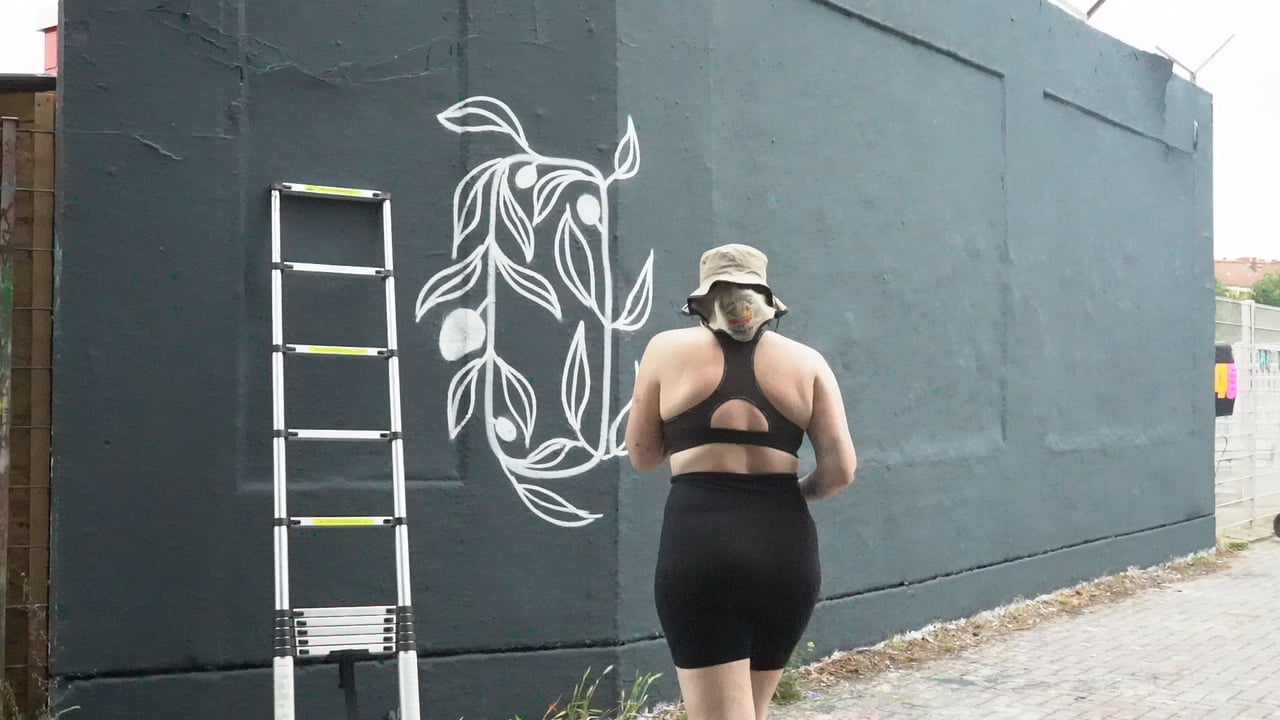 Woman in exercise attire standing in front of a large mural on a gray building wall with white line art design and a ladder nearby