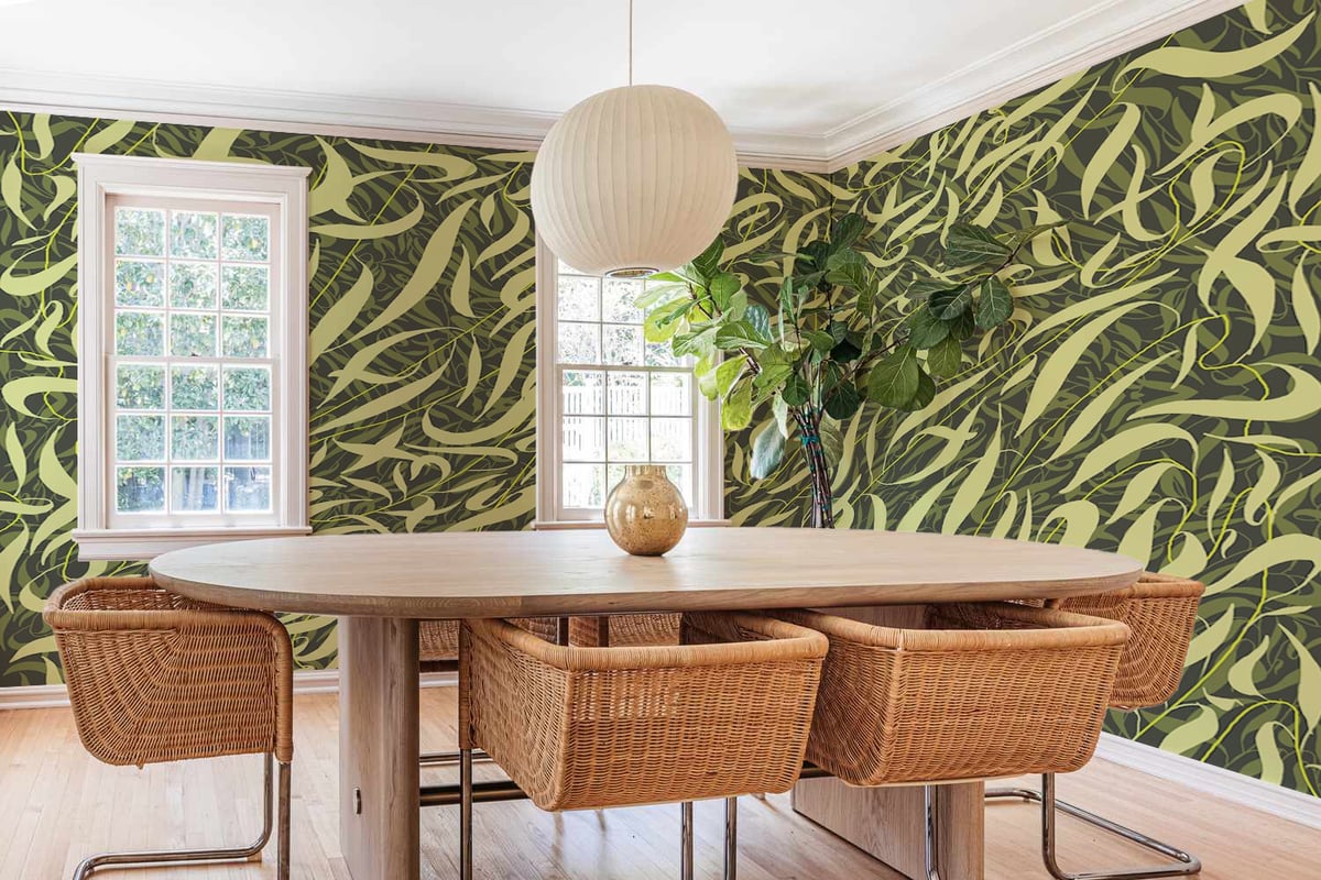 Modern dining room with bold green and yellow leaf-patterned wallpaper, wooden table, wicker chairs, pendant light, and large windows