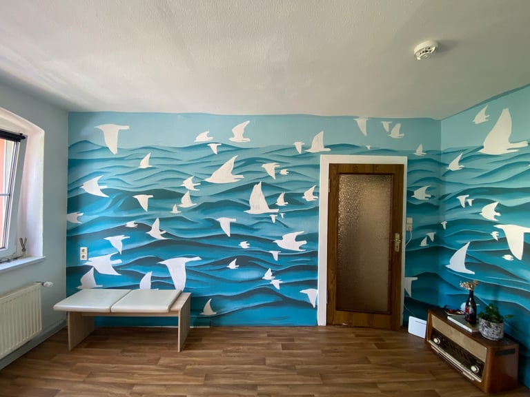 Room with large turquoise and blue mural of sailboats on wavy water covering the wall, wooden floor, white bench, and brown door
